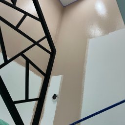 Kolorowy Kąt - Ściana w trakcie malowania na dwa kolory: beżowy u góry i biały na dole. Widoczny fragment czarnej dekoracji ściennej i niebieski pręt rusztowania.