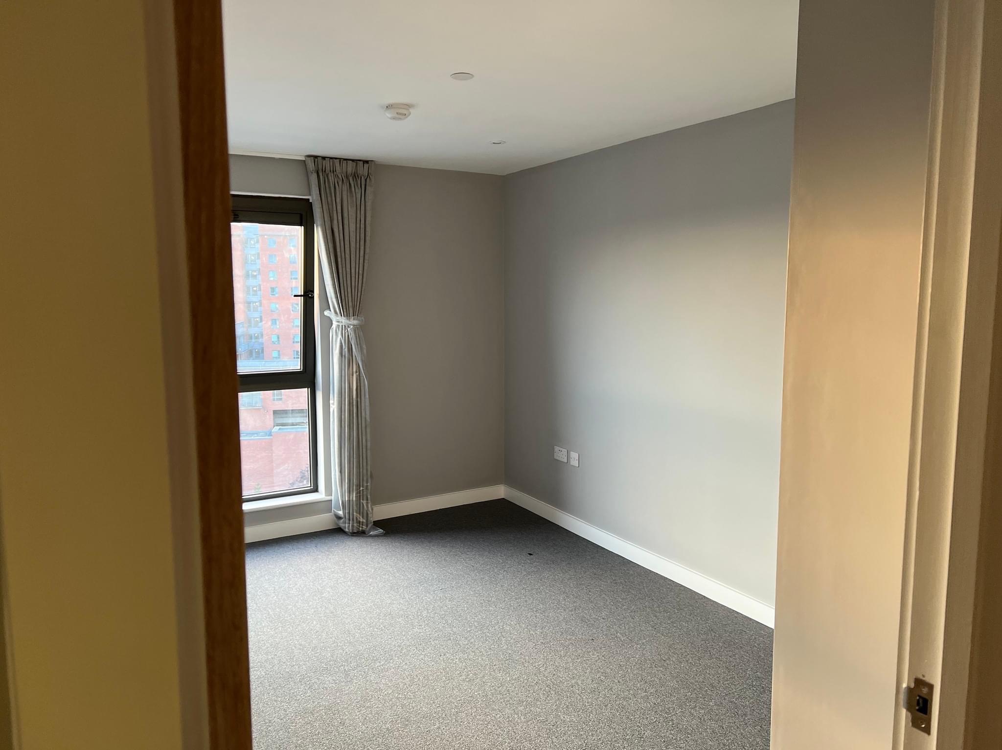 Pokój po remoncie z szarymi ścianami, szarą wykładziną i oknem z szarymi zasłonami, widok zza framugi drzwi.