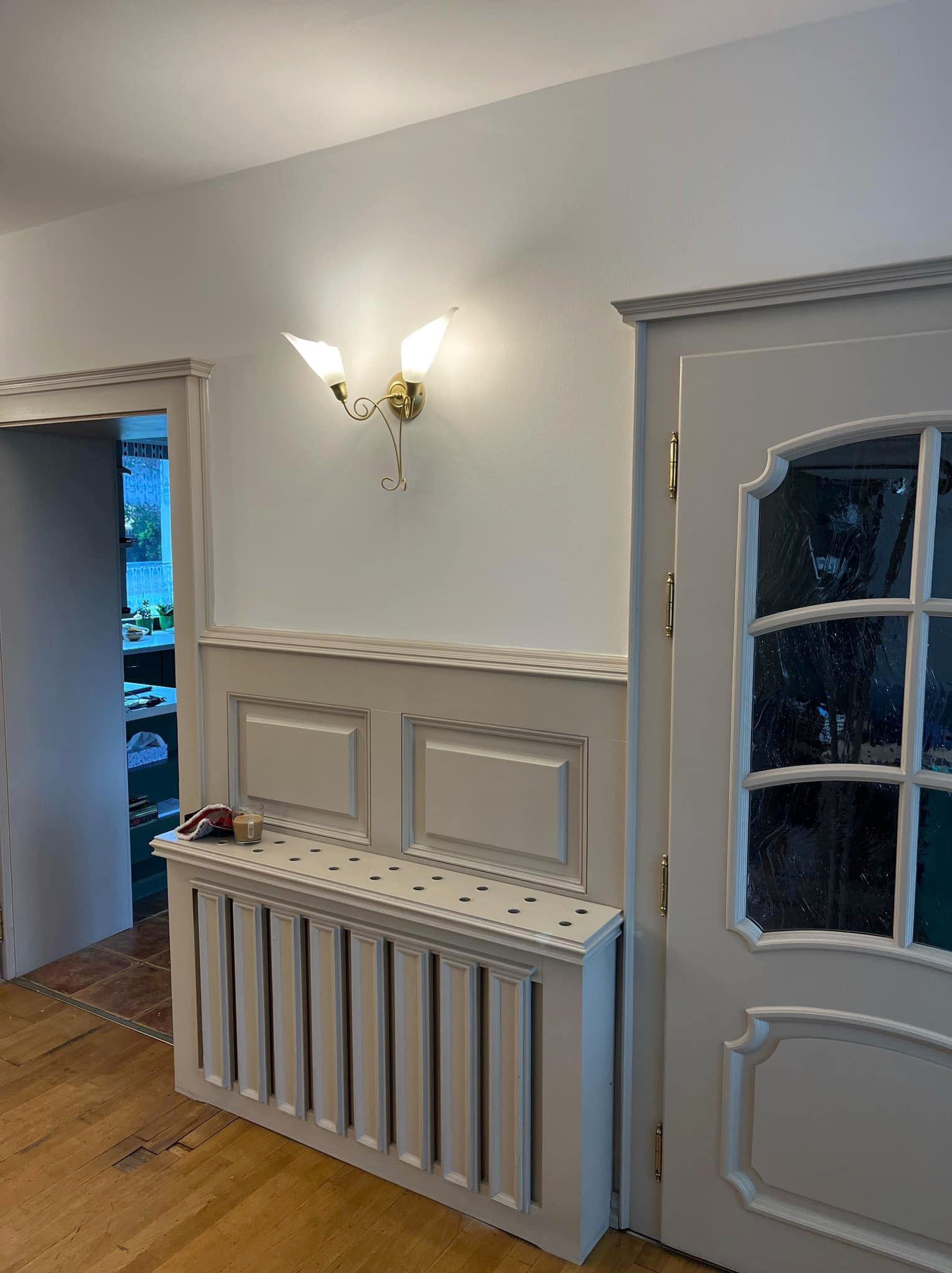 Pomalowane na jasny odcień szarości wnętrze: ściany z lameli, dekoracyjny grzejnik z ażurowym wykończeniem, drzwi z szybami i złote klamki oraz designerski kinkiet.