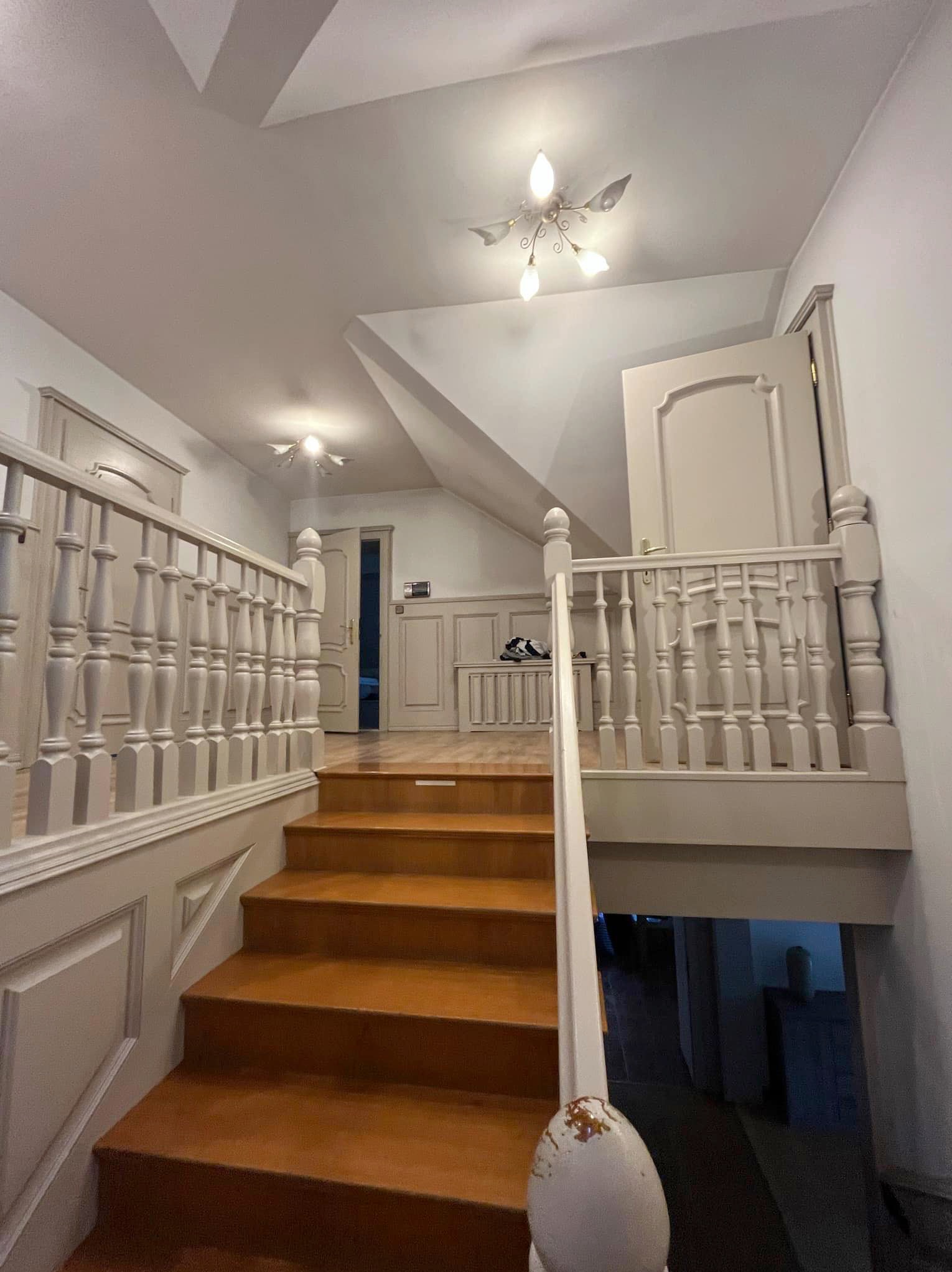 Klatka schodowa w domu jednorodzinnym z drewnianymi schodami i balustradą pomalowaną na jasny kolor, widoczne drzwi i dekoracyjne oświetlenie sufitowe.