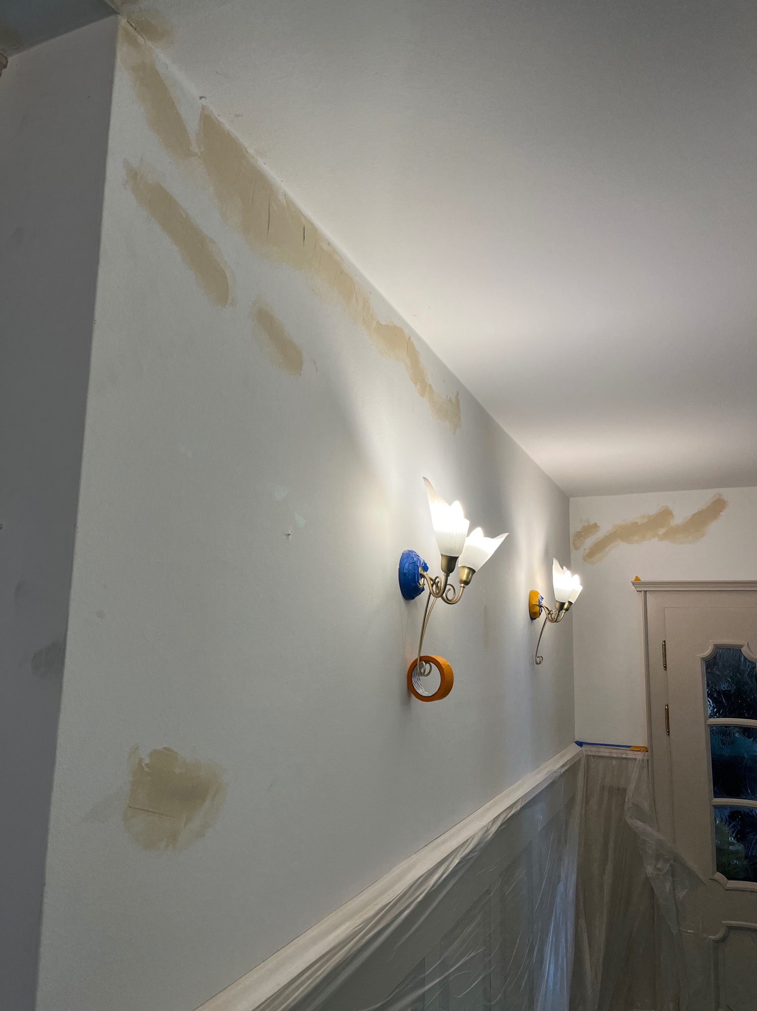 Ściana w trakcie przygotowania do malowania z zaaplikowaną warstwą wyrównującą, widoczne lampy ścienne i zabezpieczenia folią malarską.