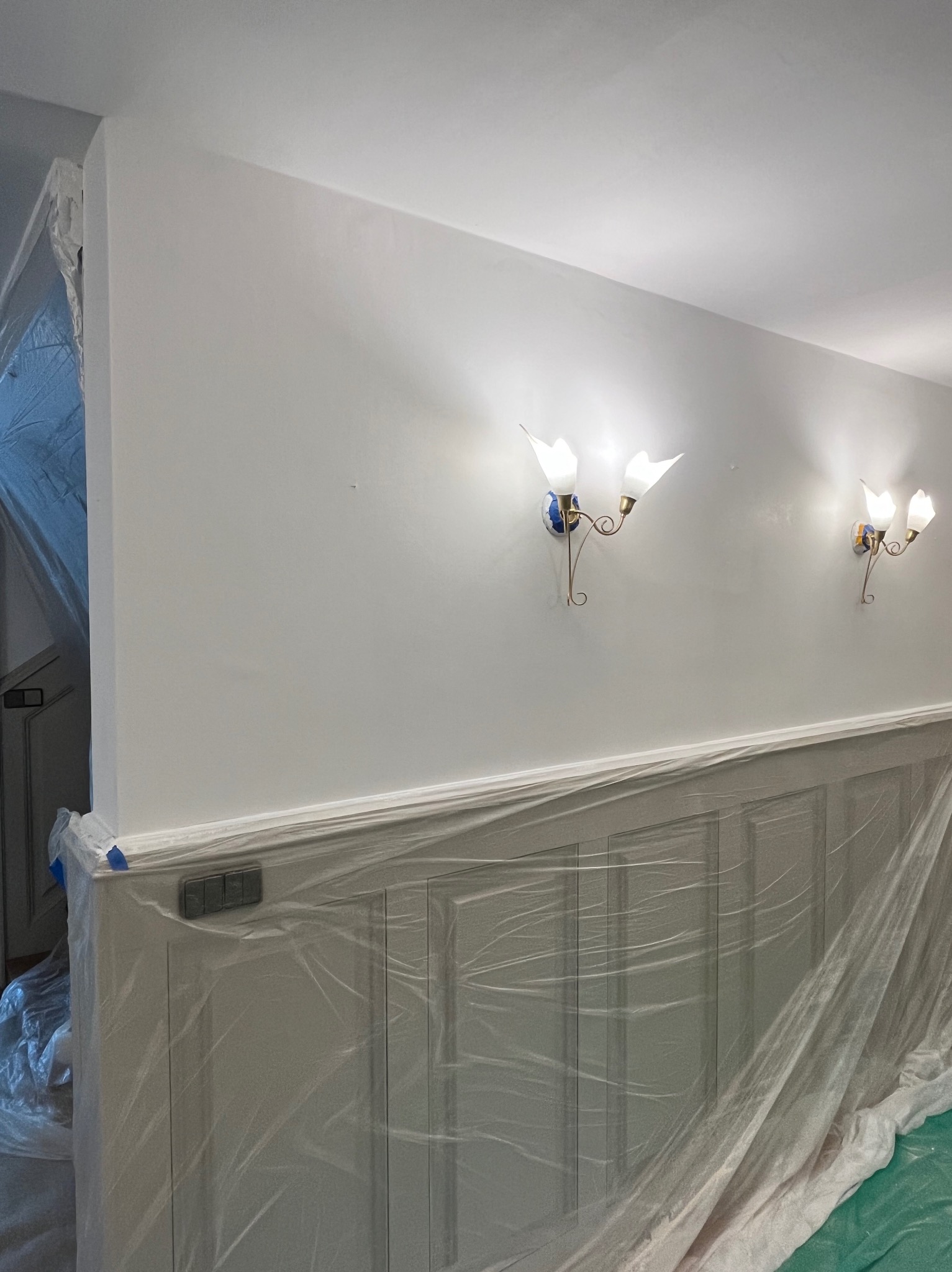 Korytarz w trakcie zabezpieczania folią malarską przed pracami wykończeniowymi. Widoczne osłonięte ściany, włączniki oraz dekoracyjne lampy ścienne.