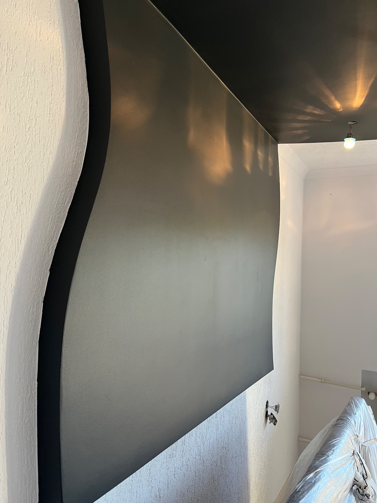 Nowoczesne wnętrze z szarą ścianą i czarnym sufitem, dekoracyjna, falista krawędź ściany, widoczne niedokończone gniazdka elektryczne i meble zabezpieczone folią.