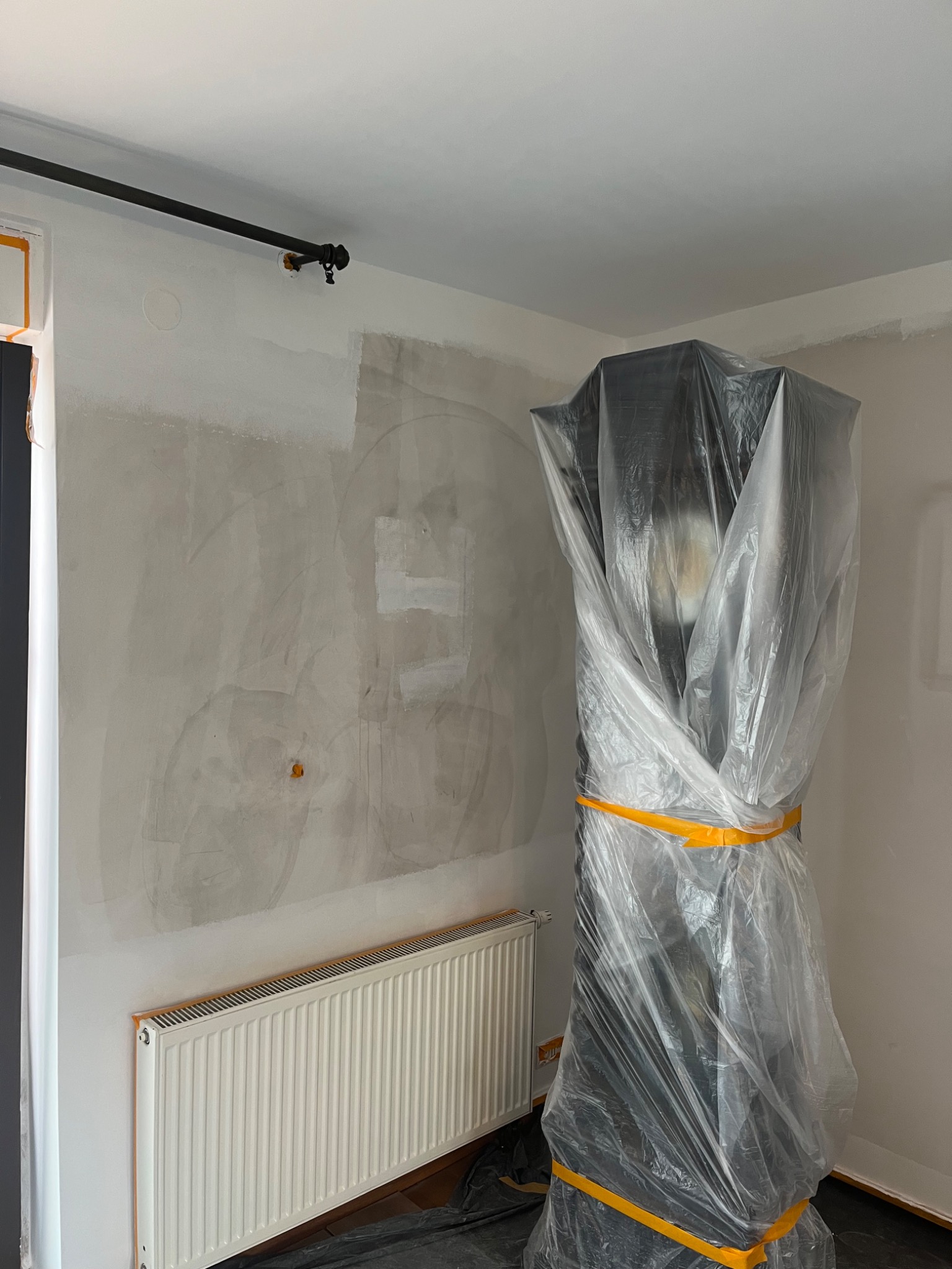 Przygotowania do prac wykończeniowych w pokoju: ściana z częściowo nałożoną warstwą wykończeniową, grzejnik i mebel zabezpieczone folią ochronną i taśmą malarską.