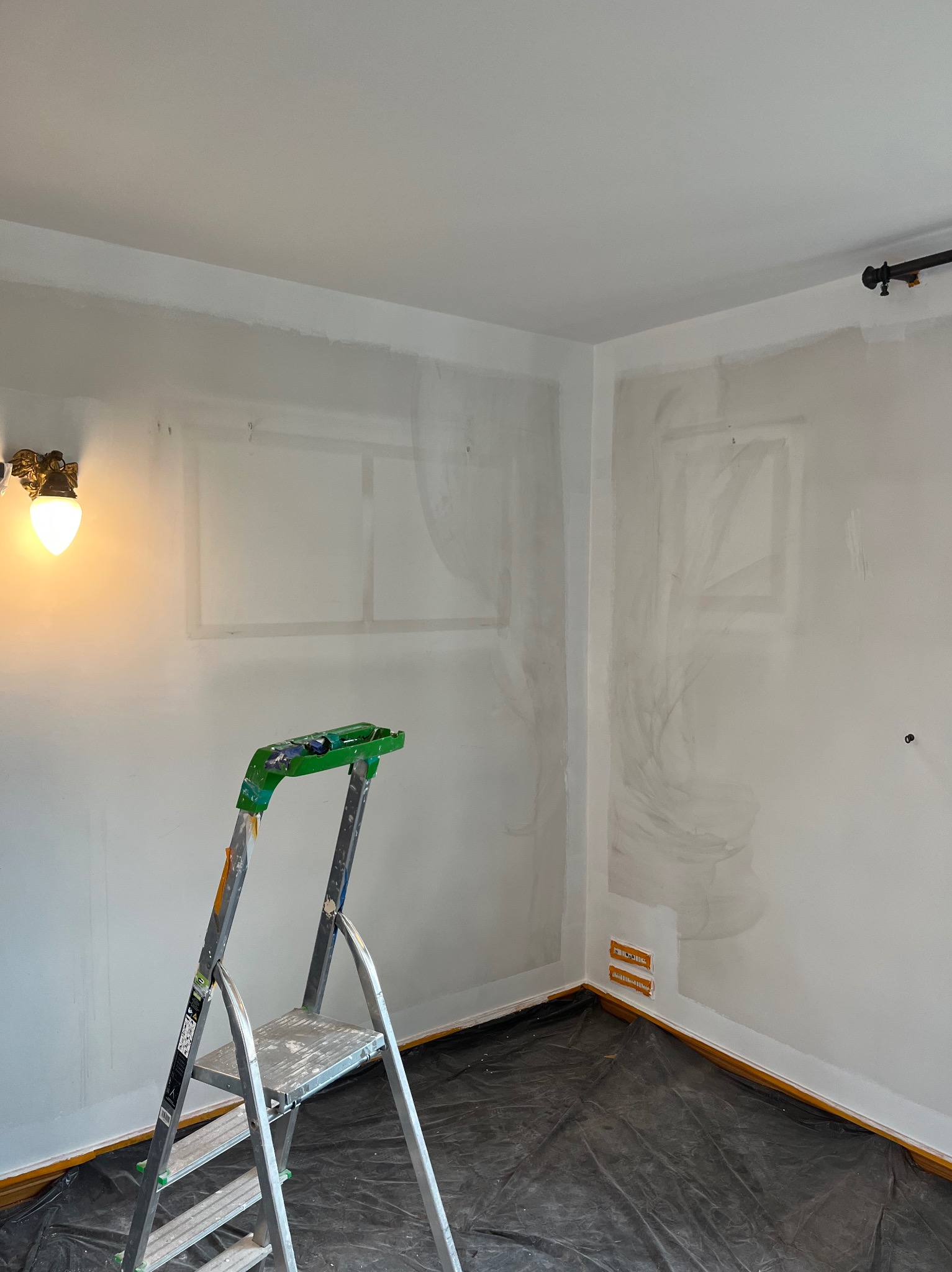 Pokój w trakcie przygotowania do malowania: ściany z śladami po szpachlowaniu, zabezpieczona podłoga folią, drabina aluminiowa z zielonymi elementami i ozdobny kinkiet z żarówką.