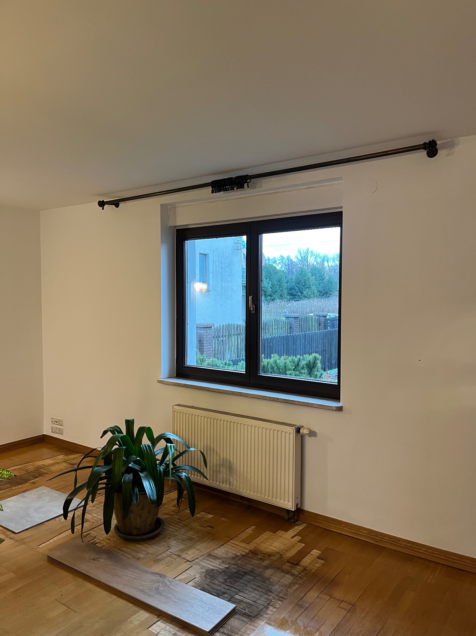 Pokój w trakcie remontu: zerwana podłoga, przygotowane panele, grzejnik pod oknem, karnisz na ścianie, roślina doniczkowa.