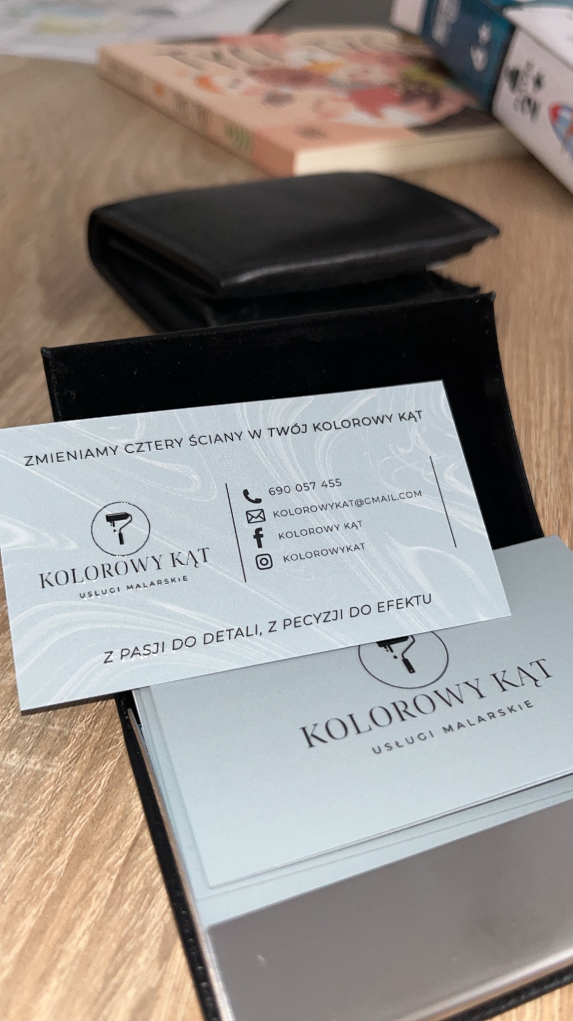 Wizytówka firmy 'Kolorowy Kąt' oferującej usługi malarskie, leżąca na drewnianym biurku, z widocznym logo pędzla i hasłem 'Zmieniamy cztery ściany w Twój kolorowy kąt'.