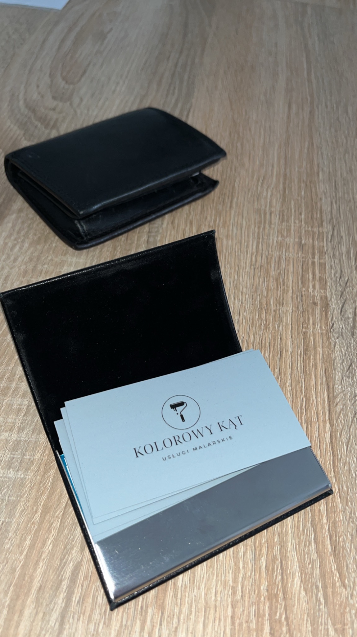 Wizytówki firmy malarskiej 'Kolorowy Kąt' w czarnym etui z metalowym wykończeniem, obok czarny portfel na drewnianym stole. Minimalistyczny design.