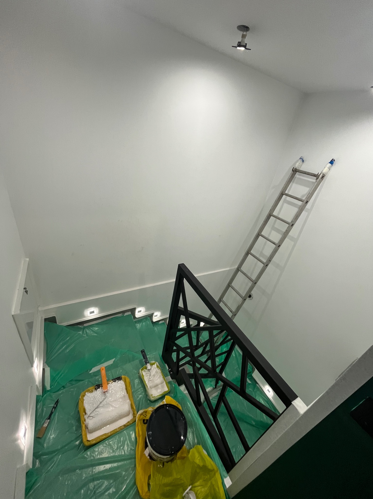 Białe ściany po malowaniu, widoczne narzędzia malarskie (wałki, kuwety) zabezpieczone folią, drabina oparta o ścianę. Ujęcie z góry, widok na klatkę schodową.