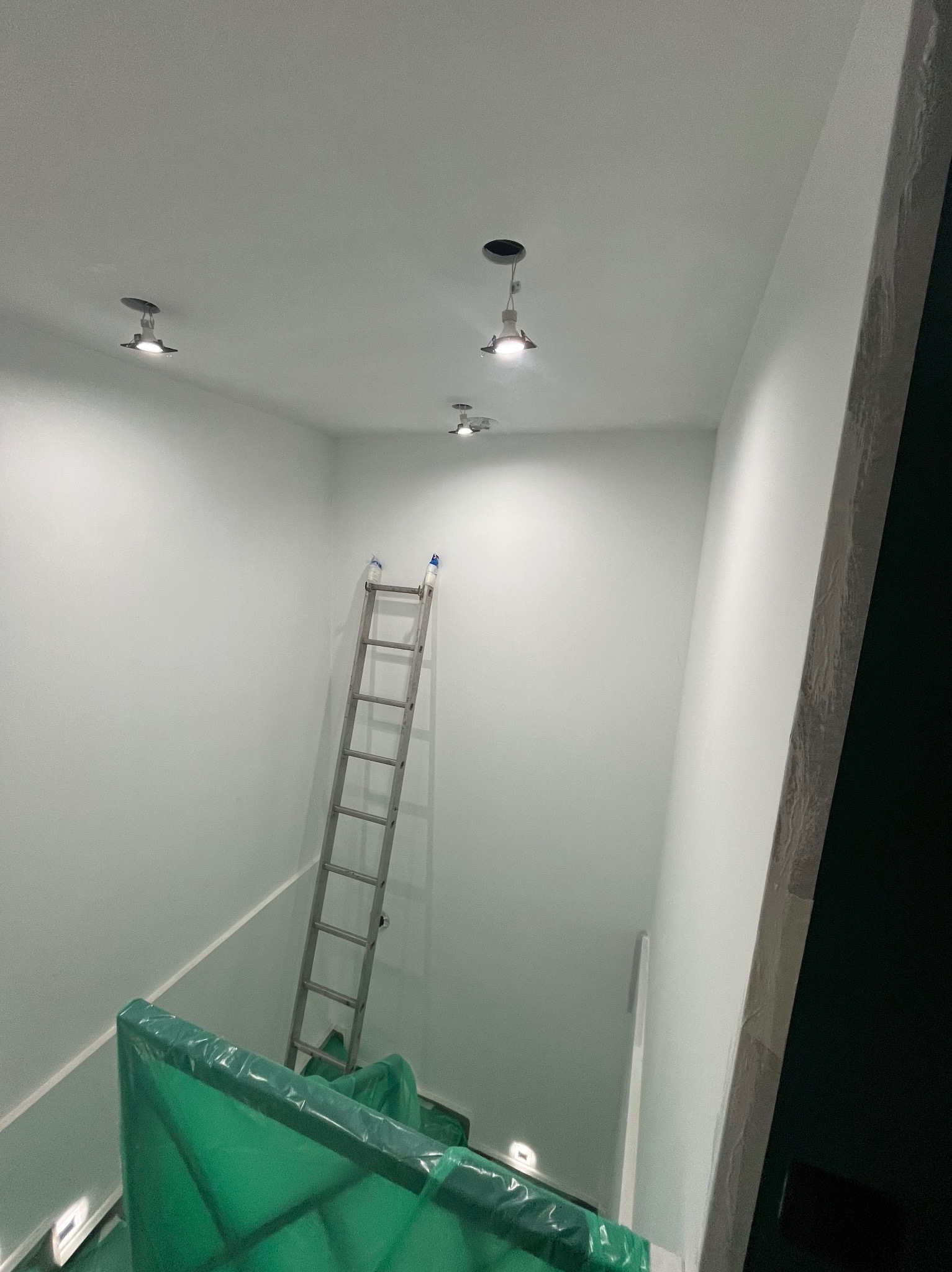 Wnętrze po szpachlowaniu: gładkie, białe ściany, drabina oparta o ścianę, zabezpieczone folią schody. Widoczne lampy sufitowe.