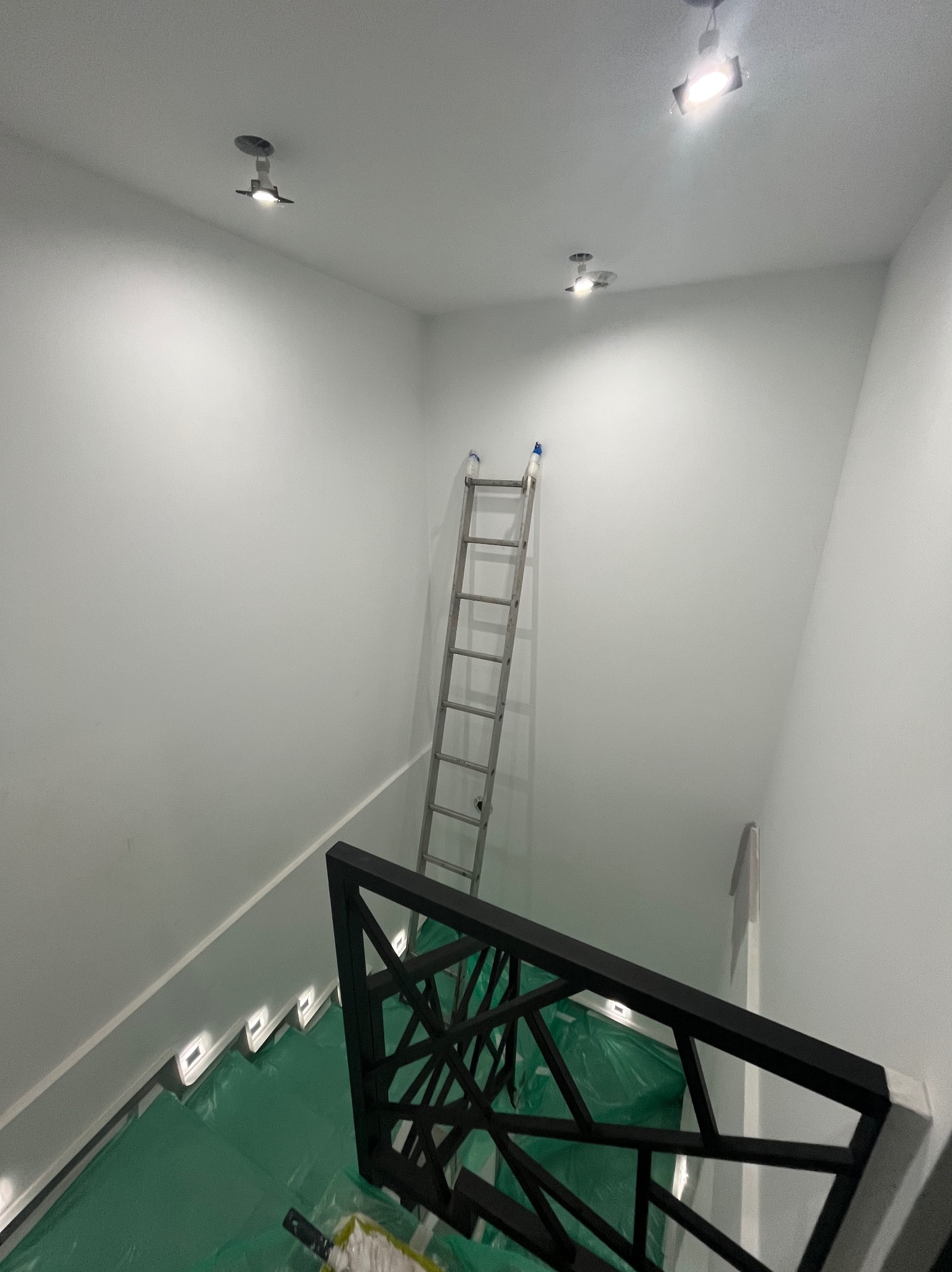 Białe ściany po malowaniu w nowoczesnym wnętrzu. Widoczna drabina oparta o ścianę, zabezpieczona podłoga i dekoracyjna balustrada schodów.