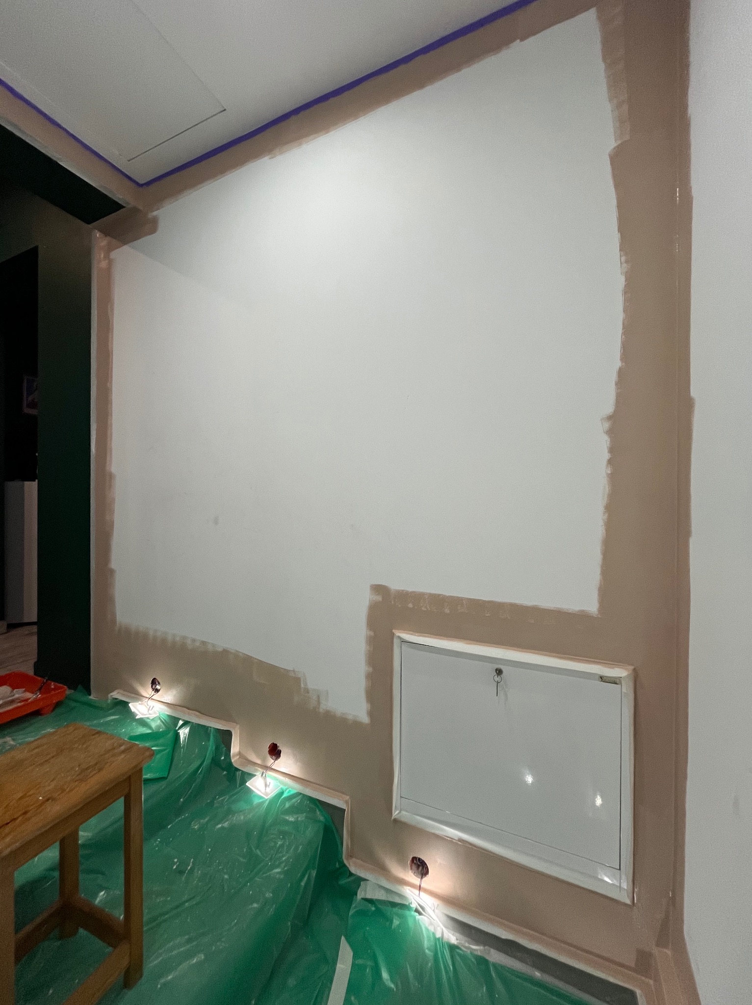 Przygotowanie do malowania: zabezpieczona folią podłoga, obklejone listwy i częściowo pomalowana ściana na biało z widocznymi śladami brązowej farby gruntującej.