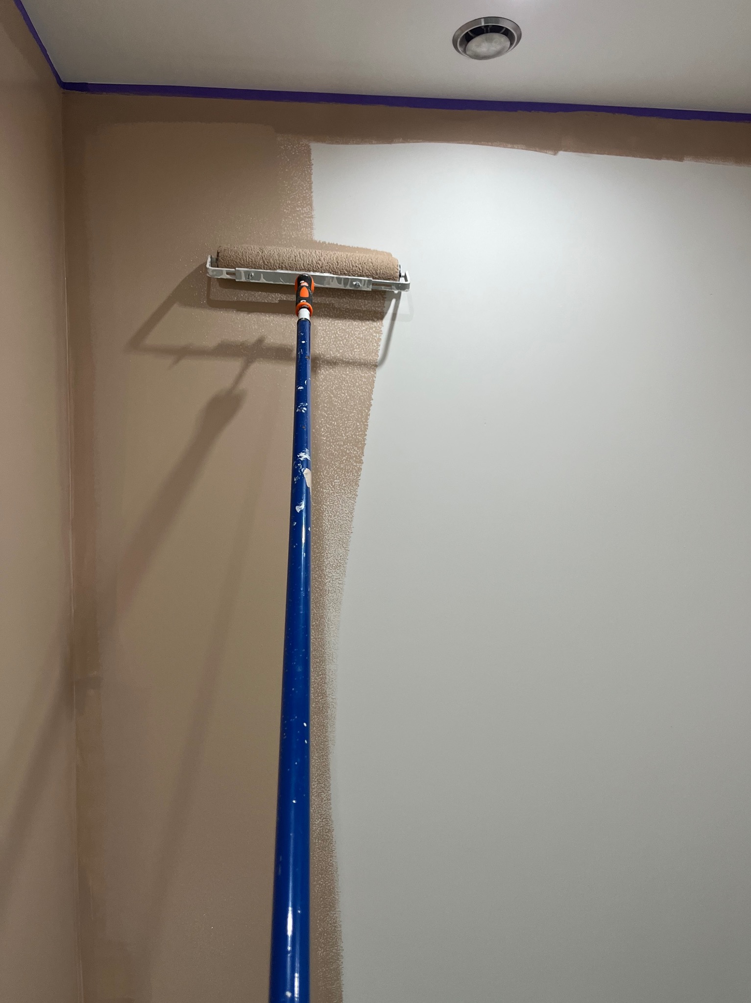 Malowanie ściany wałkiem na teleskopowym kiju. Częściowo pomalowana ściana w odcieniach beżu z niebieską taśmą malarską przy suficie.
