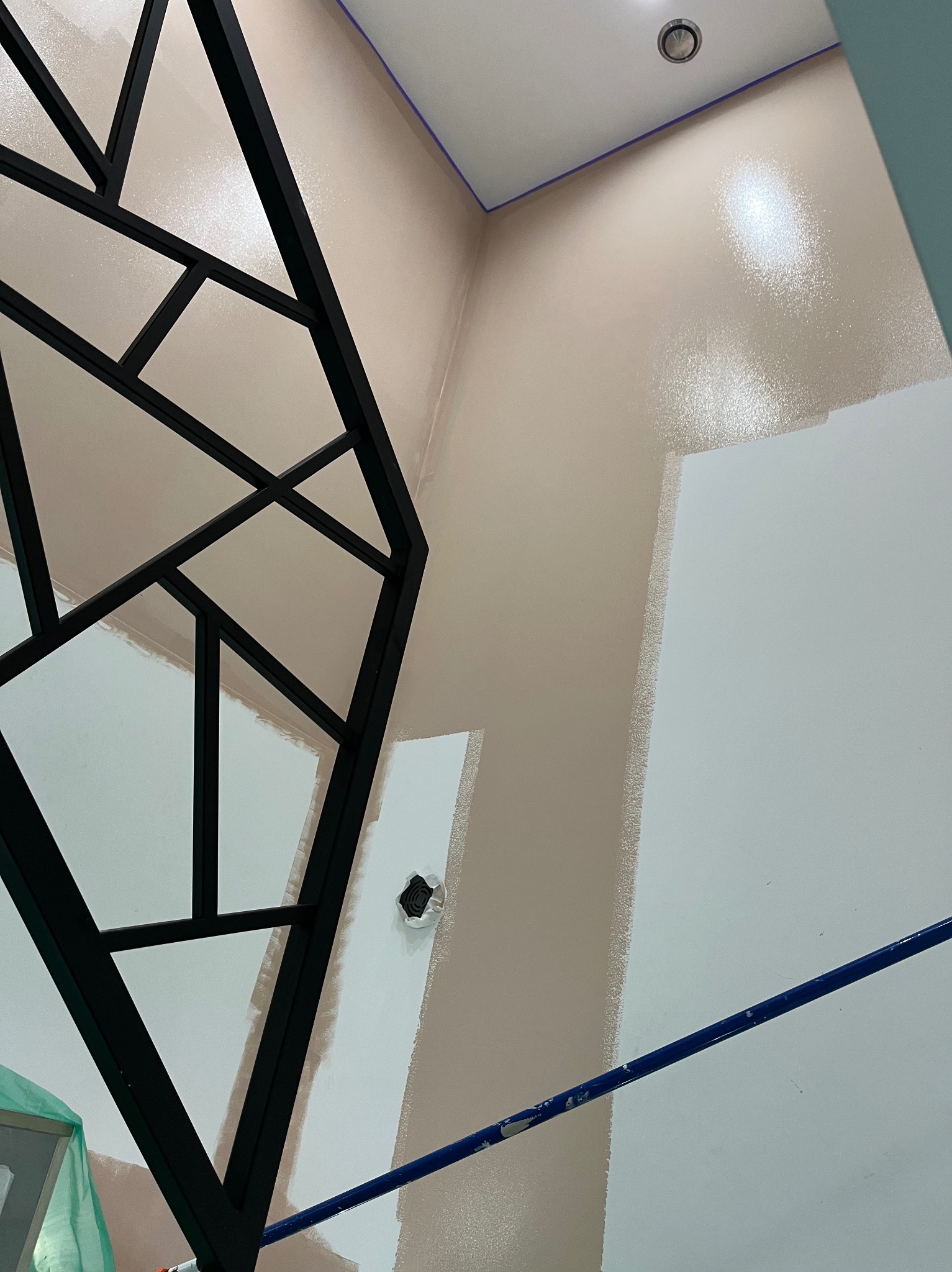 Ściana w trakcie malowania na dwa kolory: beżowy u góry i biały na dole. Widoczny fragment czarnej dekoracji ściennej i niebieski pręt rusztowania.