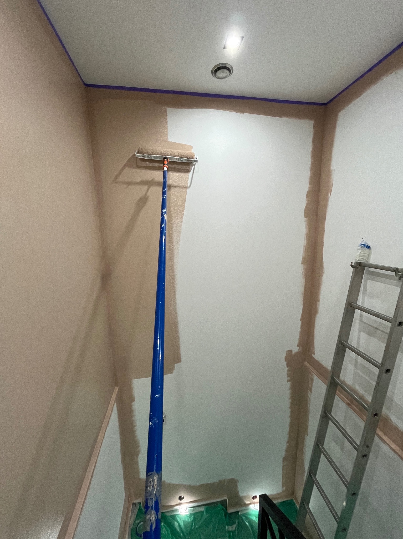 Malowanie ścian wałkiem na długim uchwycie w narożniku pomieszczenia. Widoczna drabina, taśma malarska i folia ochronna. Częściowo pomalowana ściana na biało.