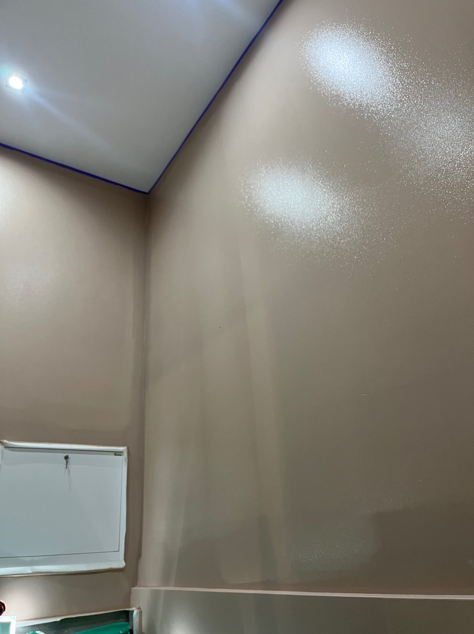 Ściana po gładzi i malowaniu na beżowo, widoczny fragment sufitu z niebieską taśmą malarską. Na ścianie widoczne refleksy światła.