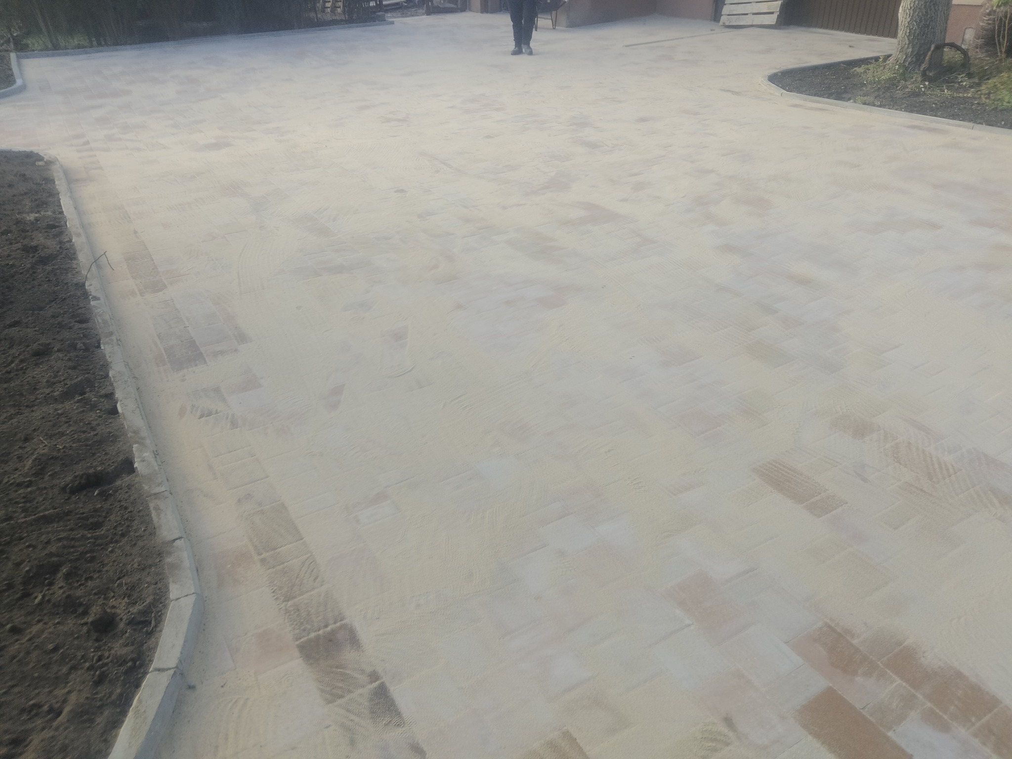 Świeżo ułożona kostka brukowa, zasypana piaskiem kwarcowym, widoczne obrzeża chodnikowe i fragment osoby w oddali.