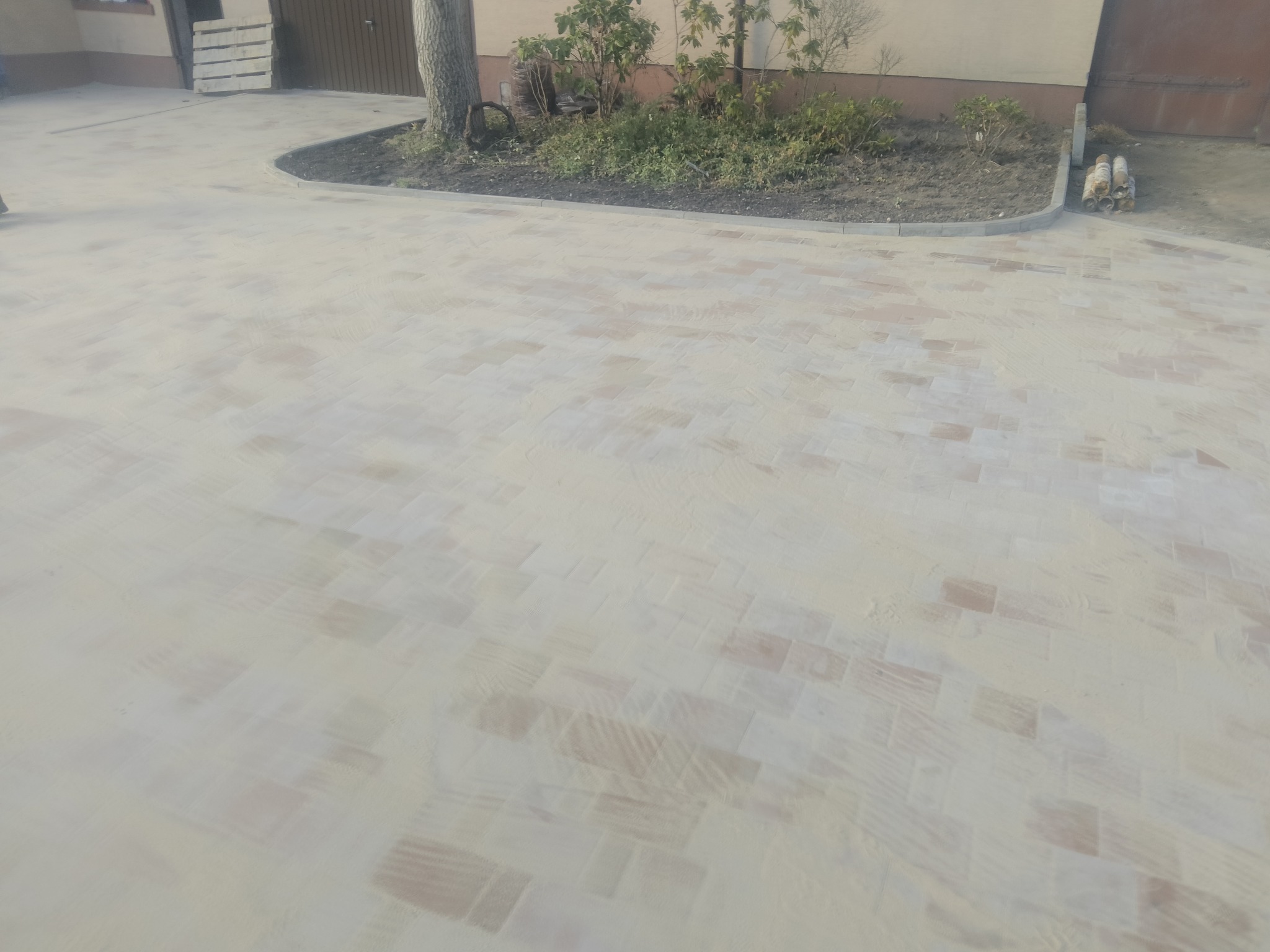 Jasna kostka brukowa ułożona na podjeździe, obrzeże z kostki oddziela rabatę z drzewami i krzewami, w tle brama garażowa i paleta.