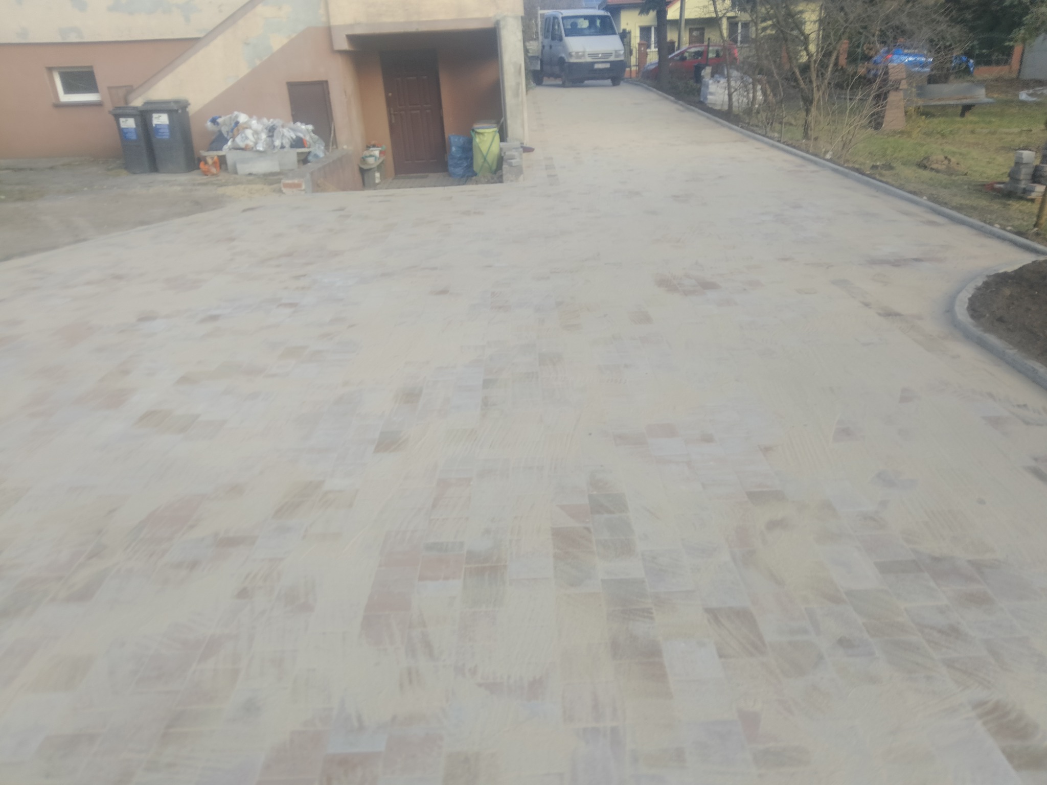 Ułożona beżowa kostka brukowa na podjeździe, widoczny fragment domu z oknem i kosze na śmieci z tyłu, oraz zaparkowany biały bus na końcu podjazdu.