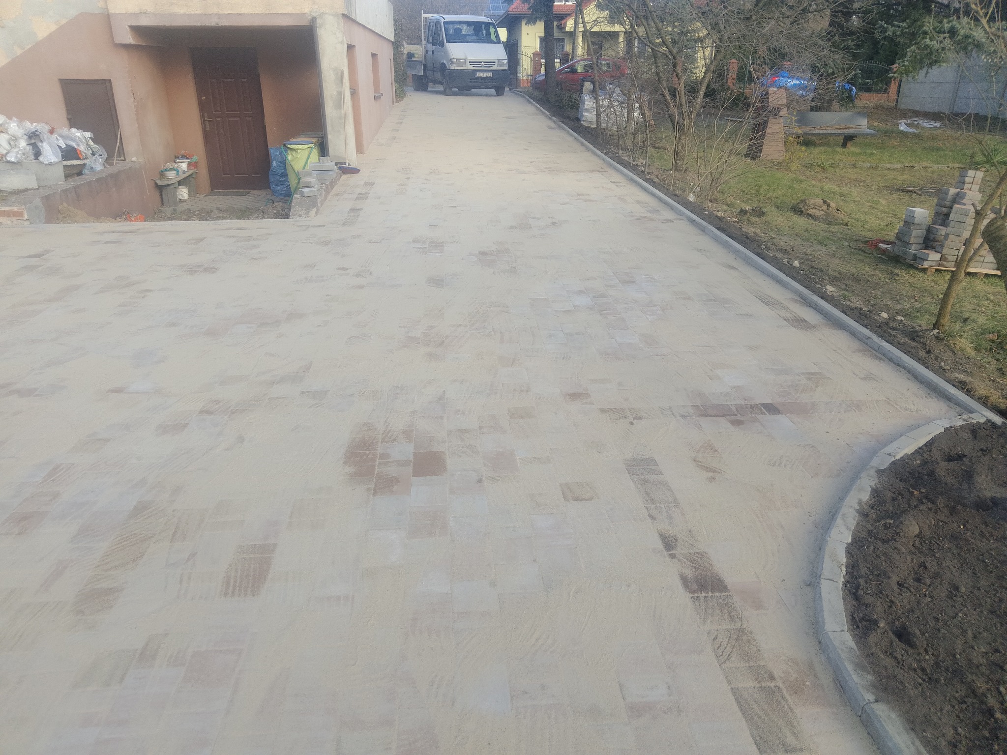 Nowo ułożona, jasna kostka brukowa na podjeździe, zasypana piaskiem, z widocznym samochodem dostawczym w tle i stosem kostki obok trawnika.