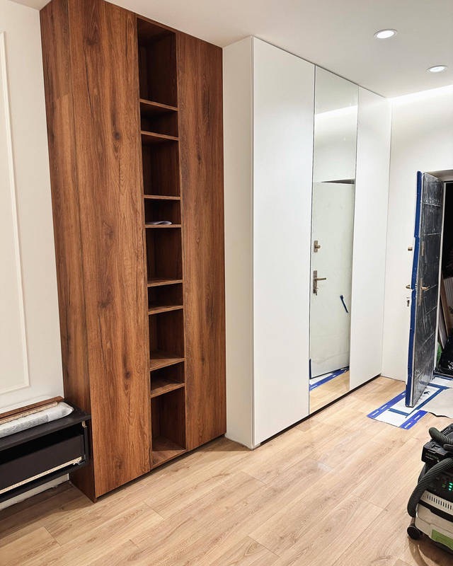 Wnętrze z zabudową: wysoka szafa z drewnianymi frontami i otwartymi półkami obok białej szafy z lustrzanymi drzwiami przesuwnymi, widoczne prace wykończeniowe.