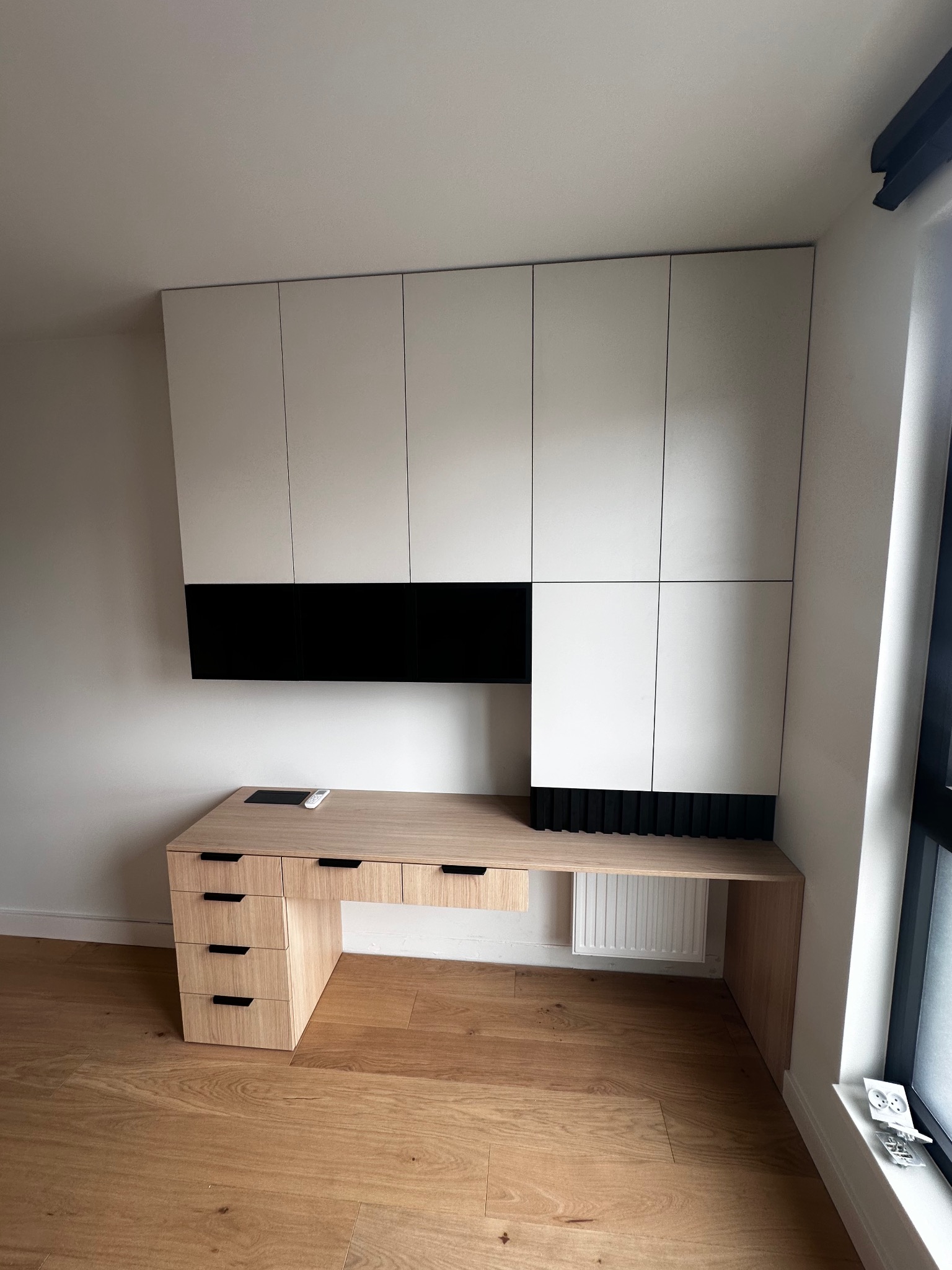 Biało-drewniana zabudowa meblowa z szafkami wiszącymi i biurkiem z szufladami, minimalistyczny design z czarnymi akcentami, widoczny fragment grzejnika i okno.