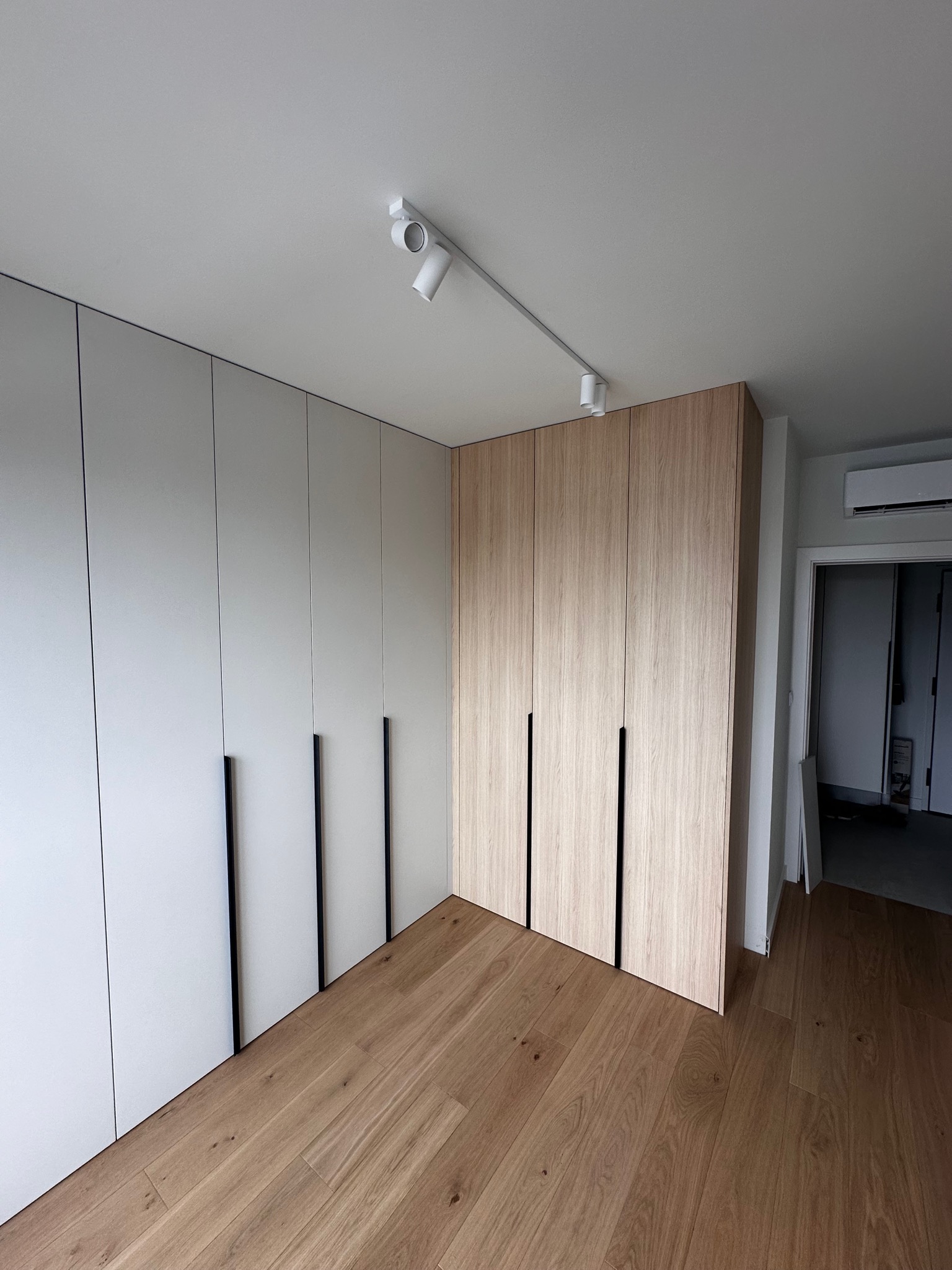 Nowoczesna szafa wnękowa z białymi i drewnianymi frontami, z czarnymi, pionowymi uchwytami, oświetlenie punktowe na suficie, drewniana podłoga. Widok z perspektywy podłogi, drzwi do innego...