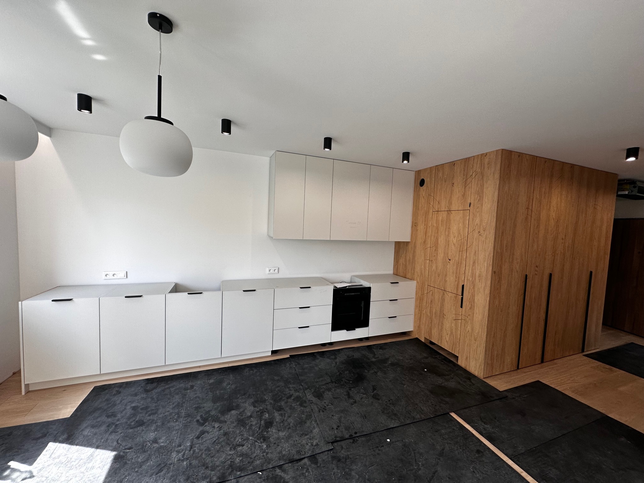 Wnętrze kuchni w trakcie montażu, widoczne białe szafki dolne i górne oraz drewniana zabudowa, podłoga zabezpieczona czarną folią.