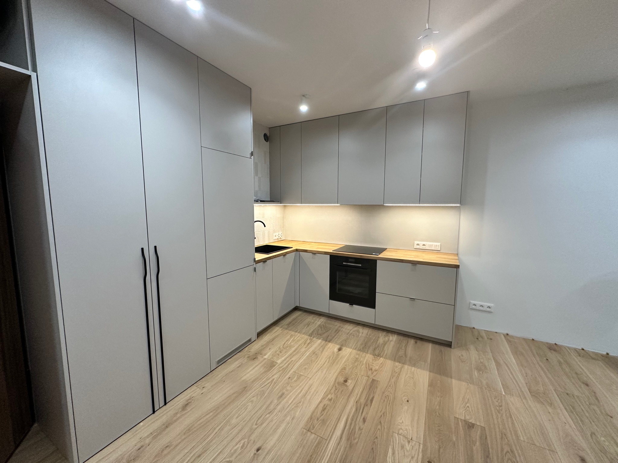 Jasnoszara kuchnia w kształcie litery L z drewnianym blatem, czarnym zlewem i piekarnikiem, widok na fragment drewnianej podłogi i białe ściany.