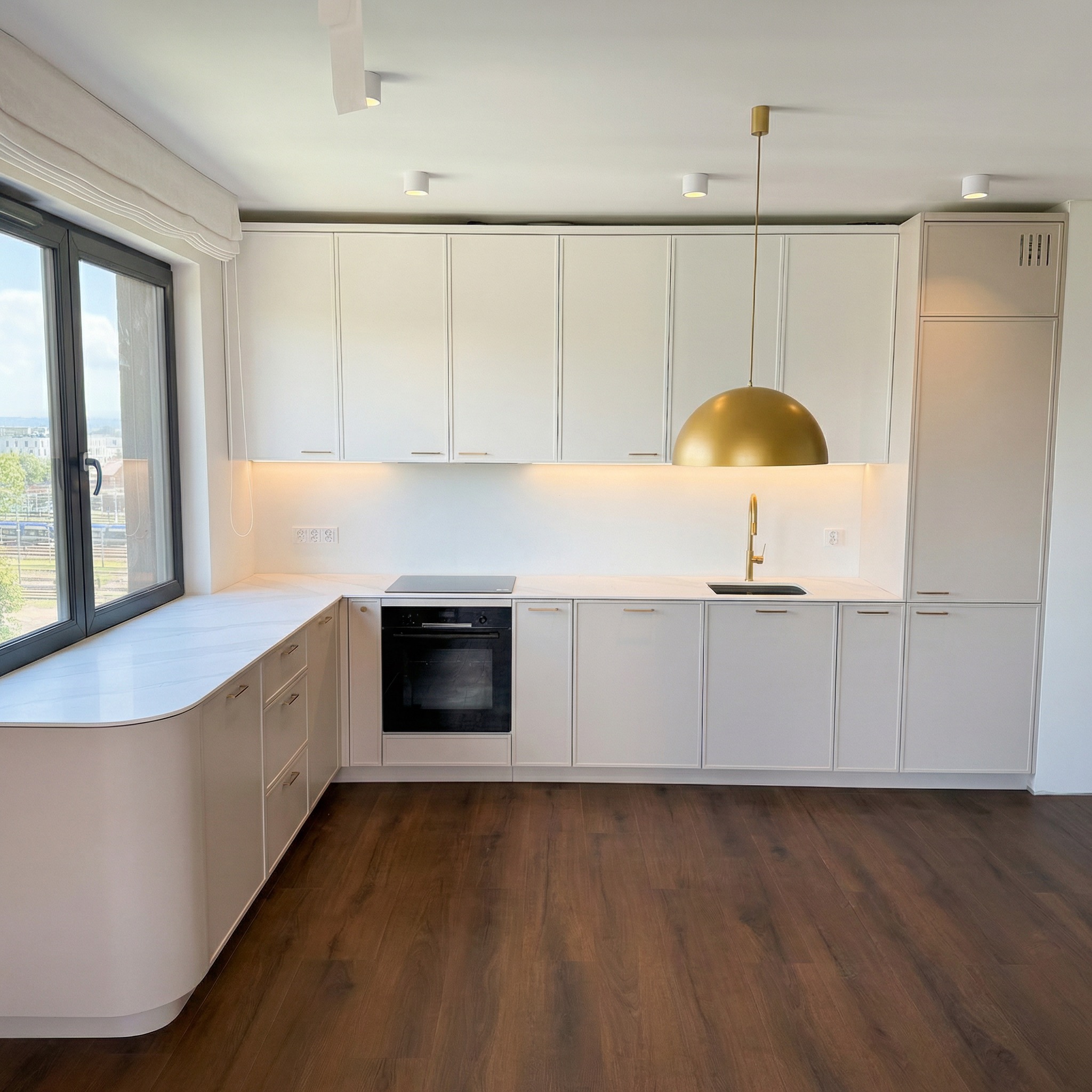 Nowoczesna, jasna kuchnia z zaokrąglonym blatem przy oknie. Złota lampa wisząca nad zlewem. Minimalistyczne fronty szafek bez uchwytów. Ciemna podłoga.