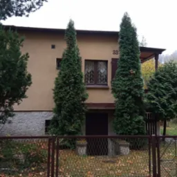 Dwukondygnacyjny dom z numerem 33, otoczony iglakami, widziany zza metalowego ogrodzenia w jesiennej scenerii.