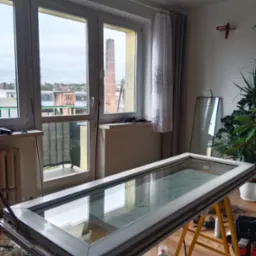 Okno PCV w trakcie naprawy na kozłach w pomieszczeniu z widokiem na komin fabryczny, widoczne narzędzia i akcesoria.