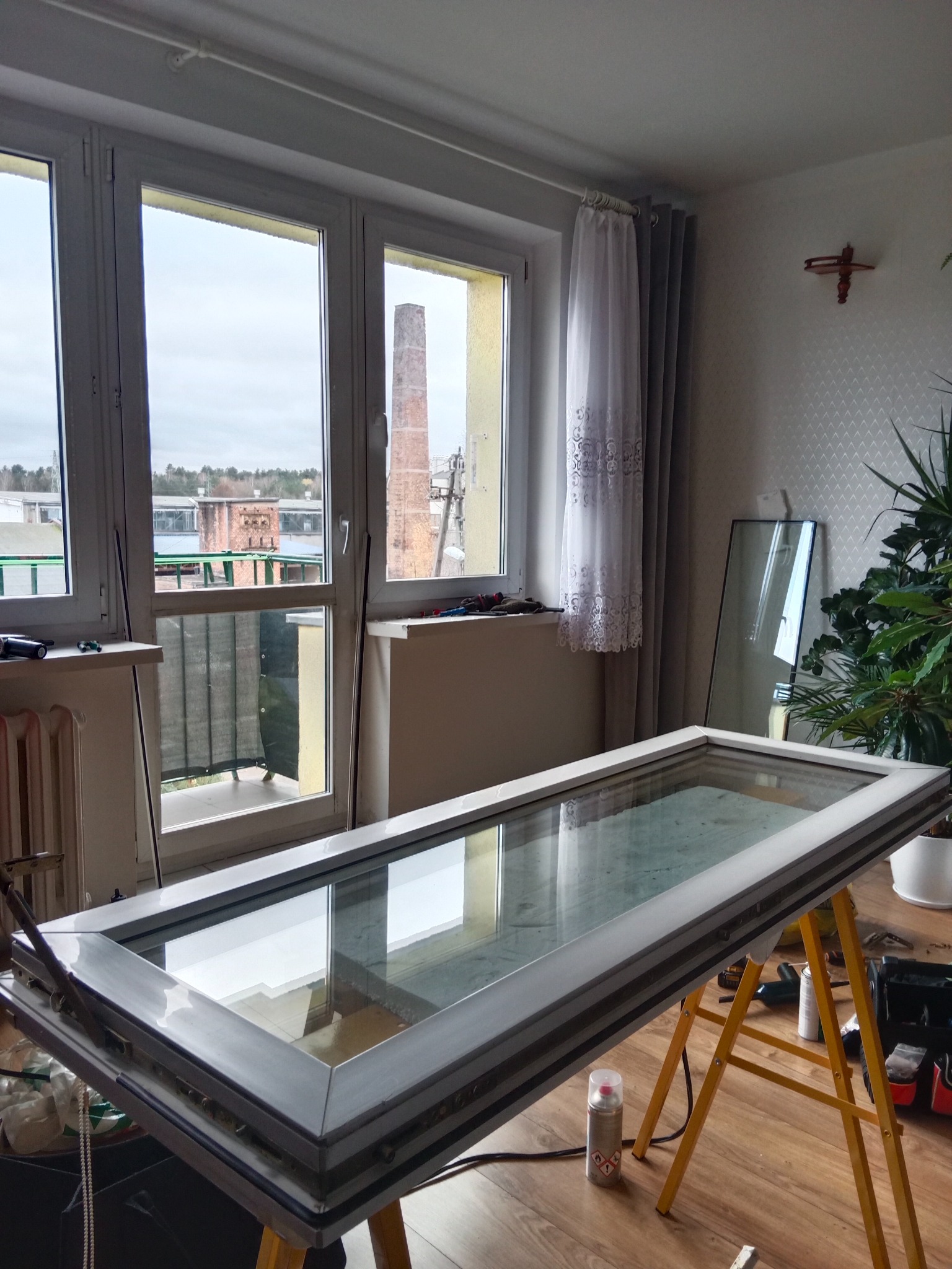 Okno PCV w trakcie naprawy na kozłach w pomieszczeniu z widokiem na komin fabryczny, widoczne narzędzia i akcesoria.