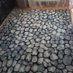 Dywany z wzorem szarych kamieni na ciemnym tle, leżący na drewnianej podłodze, widok z góry, obok szafka i zasłona okienna.