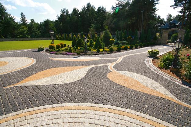 Ogród z ozdobnym podjazdem z kostki brukowej w odcieniach szarości, beżu i bieli, ułożonej we wzór przypominający liście, otoczony zielenią trawnika i drzew.