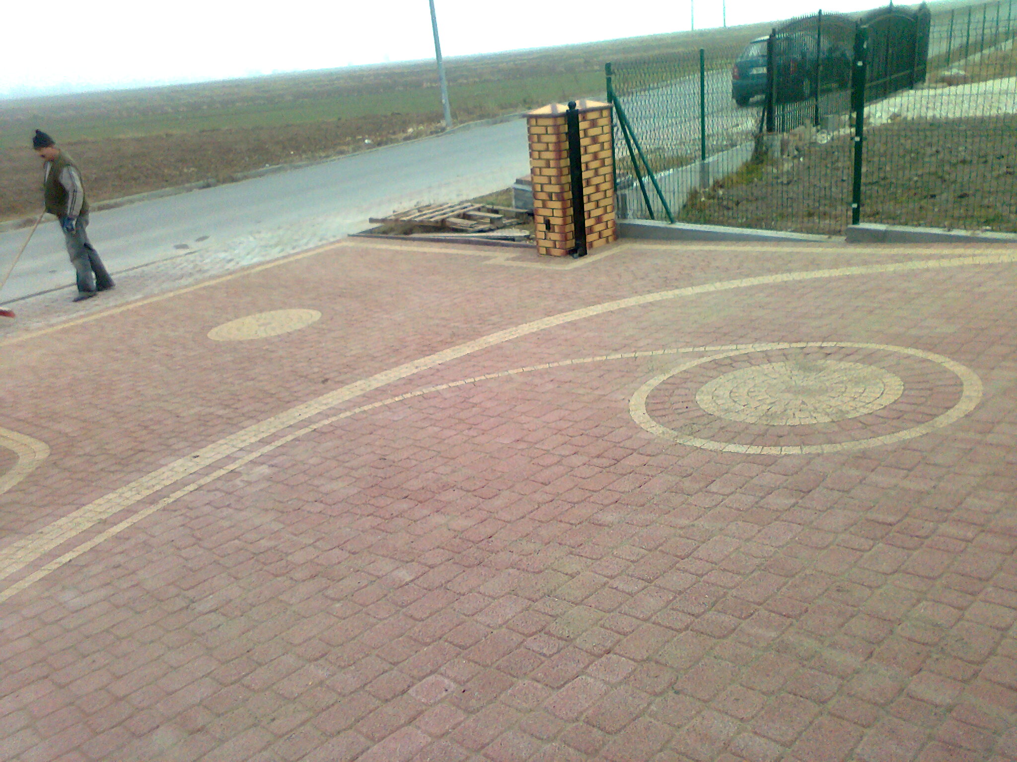 Układanie czerwonej kostki brukowej z dekoracyjnymi okręgami i pasami; pracownik zamiata powierzchnię, widoczny także element kanalizacji deszczowej z cegły klinkierowej.