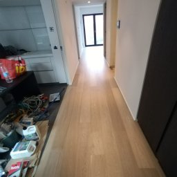 Fijas & Syn - Korytarz po remoncie z jasną drewnianą podłogą, świeżo pomalowanymi białymi ścianami i czarną zabudową meblową po prawej. W lewym dolnym rogu widoczne narzędzia i materiały malarskie, sugerujące...
