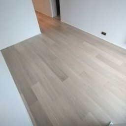 Fijas & Syn - Jasna, drewniana podłoga ułożona w jodełkę, świeżo po remoncie, z białymi ścianami i widocznymi gniazdkami elektrycznymi. W tle fragment otwartych drzwi.