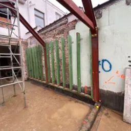 Konstrukcja nośna z belek stalowych i drewnianych słupków, przygotowana do murowania ściany na zewnątrz budynku z widocznymi elementami rusztowania i oznaczeniami na ścianie.