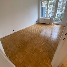 Fijas & Syn - Pusty pokój z parkietem w jodełkę, świeżo pomalowanymi białymi ścianami i grzejnikiem pod oknem balkonowym z widokiem na budynek mieszkalny.