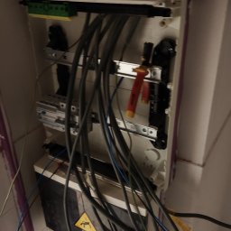 Fijas & Syn - Otwarta skrzynka elektryczna z licznymi kablami, zabezpieczeniami i narzędziami, widoczny znak ostrzegawczy o ryzyku porażenia prądem, instalacja w trakcie prac.