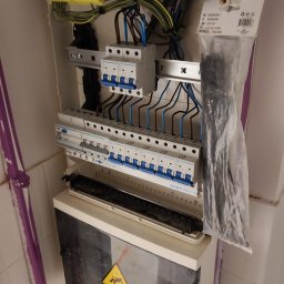 Fijas & Syn - Otwarta skrzynka rozdzielcza z zabezpieczeniami, przewodami, listwami zaciskowymi i paczką opasek kablowych, zamontowana na białej ścianie z fioletowymi elementami dekoracyjnymi.