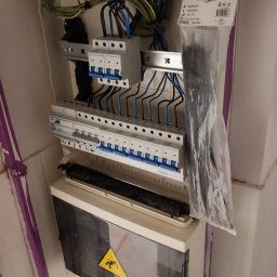 Fijas & Syn - Otwarta skrzynka elektryczna z bezpiecznikami, przewodami i listwą zaciskową, z plastikowym workiem z opaskami kablowymi obok, oraz żółtym znakiem ostrzegawczym na dolnej pokrywie, montowana...