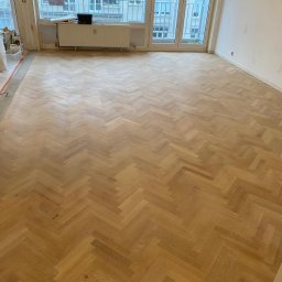 Fijas & Syn - Ułożony parkiet w jodełkę w trakcie renowacji, zabezpieczony folią przy ścianach, widoczne ślady szpachlowania na ścianie, okno balkonowe z widokiem na miasto.