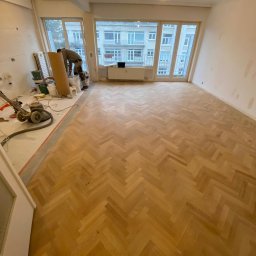 Fijas & Syn - Parkiet ułożony w jodełkę w trakcie renowacji, widoczna maszyna do cyklinowania i materiały do wykończenia podłogi, ściany przygotowane do malowania, duże okno z widokiem na budynek mieszkalny.