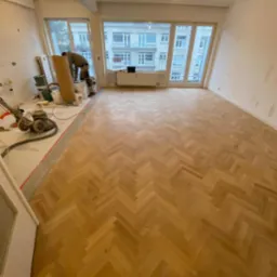 Parkiet ułożony w jodełkę w trakcie renowacji, widoczna maszyna do cyklinowania i materiały do wykończenia podłogi, ściany przygotowane do malowania, duże okno z widokiem na budynek mieszkalny.