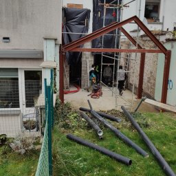 Fijas & Syn - Konstrukcja stalowa dobudowy domu z widocznymi rusztowaniami, częściowo otynkowaną ścianą i rurami na trawie.