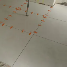 Układanie jasnych płytek podłogowych z użyciem pomarańczowych klipsów poziomujących. W tle widoczne kable elektryczne i surowe ściany w trakcie remontu.