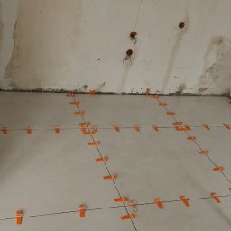 Fijas & Syn - Układanie jasnych płytek podłogowych z użyciem pomarańczowych krzyżyków dystansowych i czarnych sznurków. Widoczne wyprowadzenia elektryczne na ścianie.