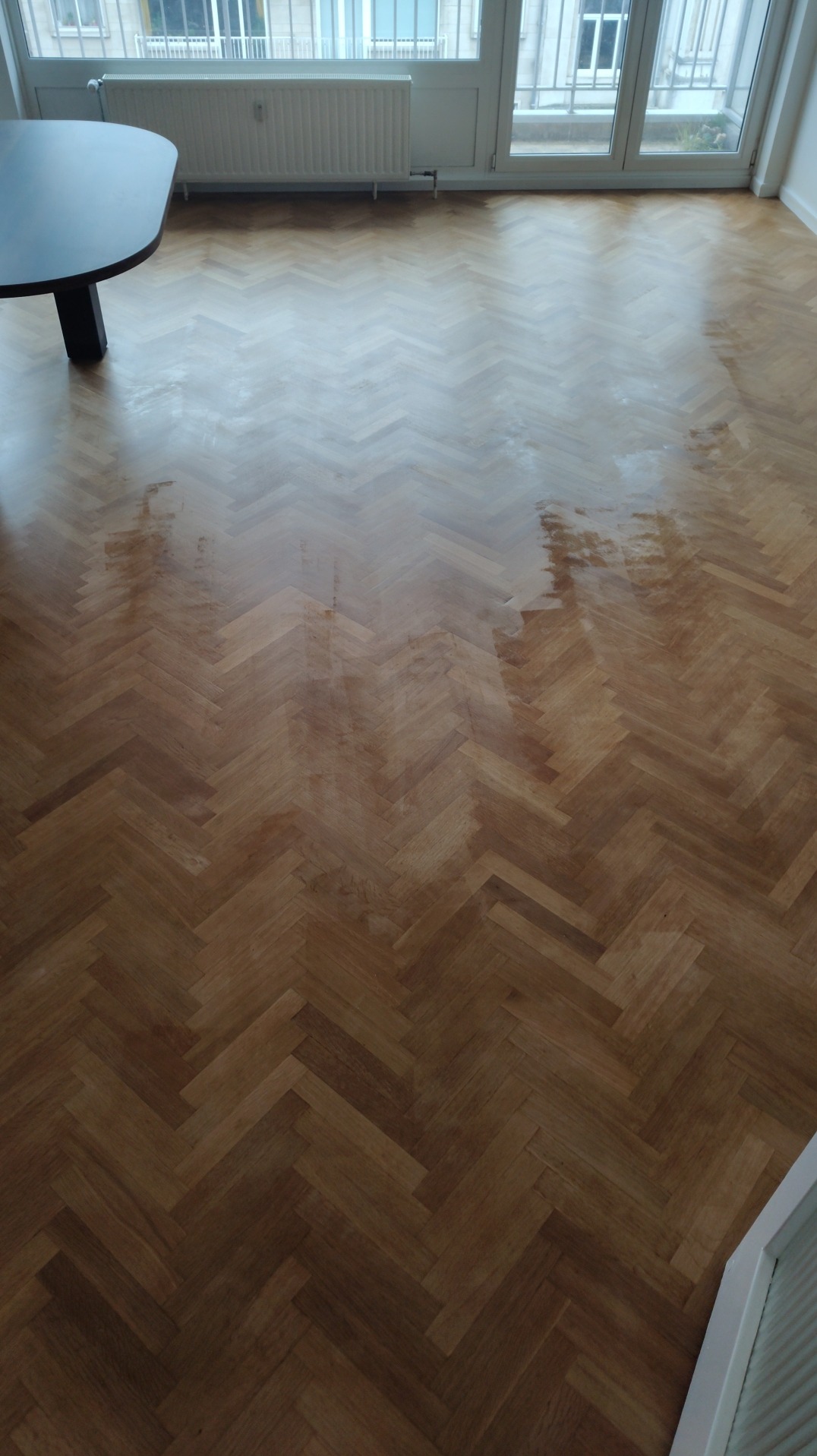 Parkiet ułożony w jodełkę, częściowo świeżo polakierowany, widoczne jaśniejsze i ciemniejsze fragmenty, stół z czarnym blatem w lewym górnym rogu, okno balkonowe z grzejnikiem.